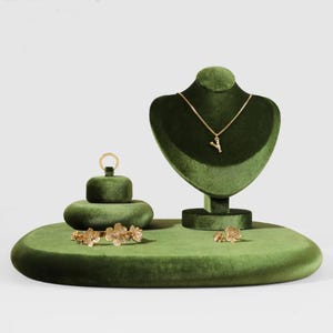 May include: A green velvet jewelry display set. The set includes a necklace bust, a small ring holder, and an oval base. A gold necklace with a letter pendant, a pair of earrings, and a floral bracelet are displayed on the set.