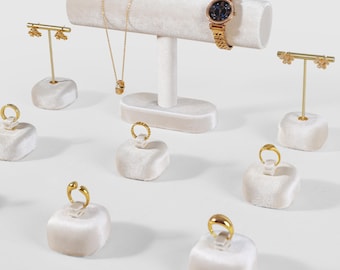 Puede incluir: Exhibición de joyas sobre soportes de terciopelo color crema. Un reloj de oro, un collar y pendientes se exhiben en un soporte en T. Varios anillos de oro se muestran en soportes individuales de terciopelo. Fondo blanco.
