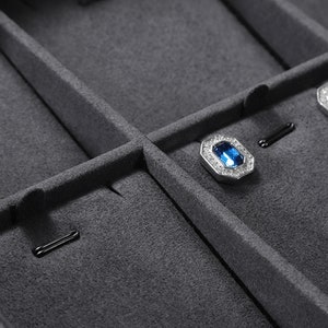Puede incluir: Primer plano de una caja de joyer&iacute;a de terciopelo gris con compartimentos para guardar pendientes y otras joyas peque&ntilde;as. La caja tiene un forro de terciopelo negro y un cierre de metal plateado.
