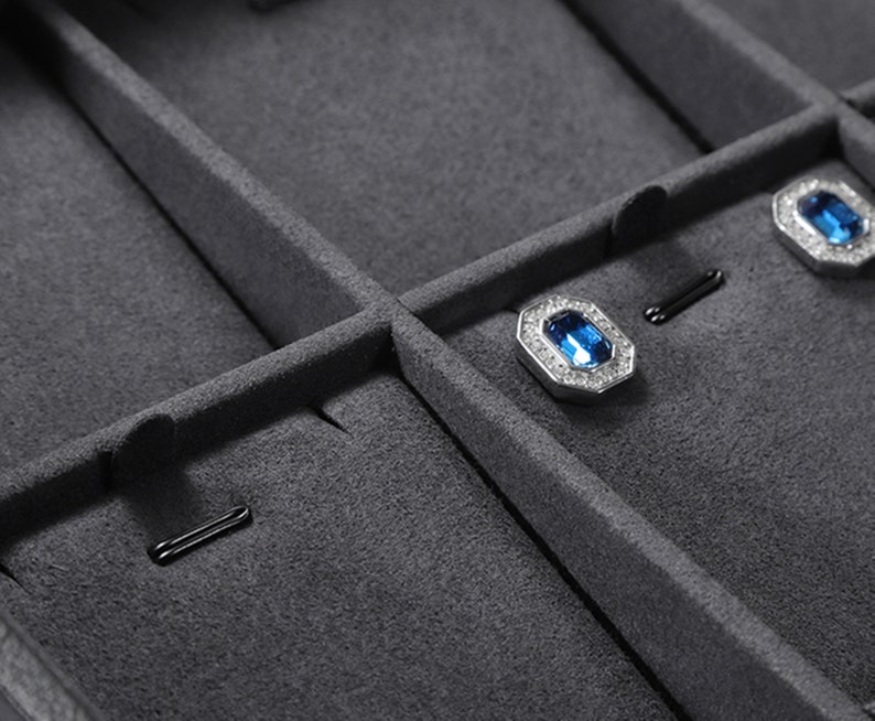 Puede incluir: Primer plano de una caja de joyer&iacute;a de terciopelo gris con compartimentos para guardar pendientes y otras joyas peque&ntilde;as. La caja tiene un forro de terciopelo negro y un cierre de metal plateado.