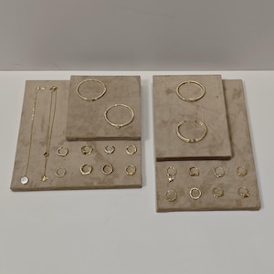 以下が含まれることがあります： ベージュのベルベットで覆われた2つのボードに展示されたゴールドトーンのジュエリーコレクション。2本の繊細なネックレス、2本の細いブレスレット、1本のオープンカフブレスレット、そして様々なデザインとサイズの16個の小さなフープピアスが含まれています。