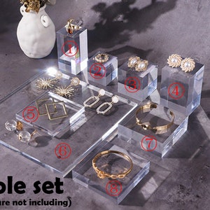 May include: A set of eight clear acrylic display stands for jewelry. The stands are in various sizes and shapes, including square, rectangular, and round. The stands are perfect for displaying earrings, necklaces, bracelets, and rings.
