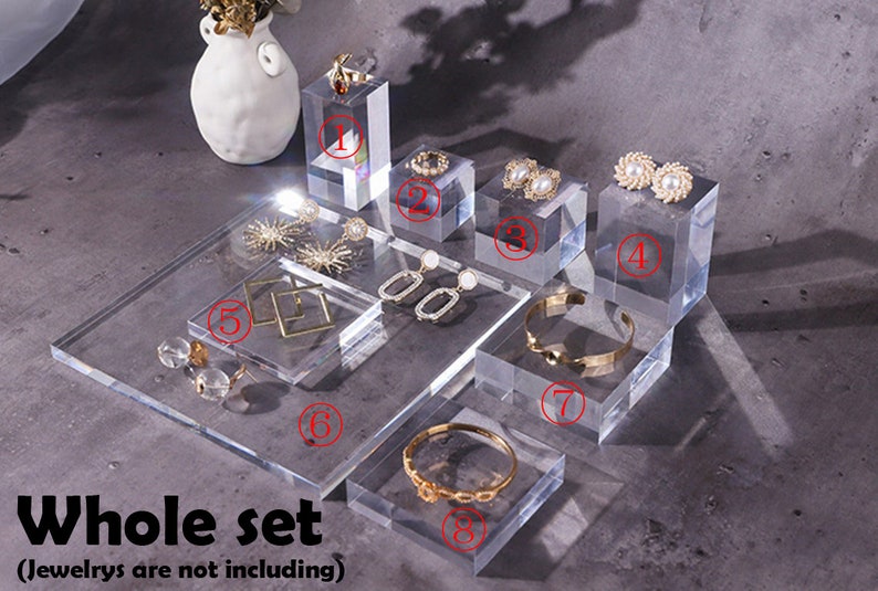May include: A set of eight clear acrylic display stands for jewelry. The stands are in various sizes and shapes, including square, rectangular, and round. The stands are perfect for displaying earrings, necklaces, bracelets, and rings.