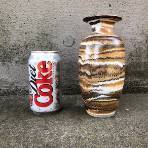 May include: A tall, ceramic vase with a wide base and a flared rim. The vase features horizontal bands of brown, white, and tan colors. A Diet Coke can is next to the vase for size comparison.