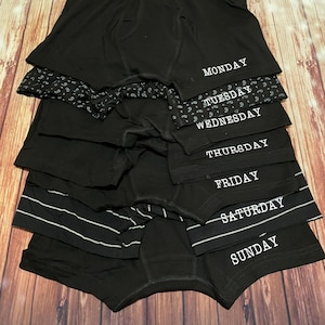 May include: A stack of seven black boxer briefs with white waistband and white text. The text on each pair reads: Monday, Tuesday, Wednesday, Thursday, Friday, Saturday, Sunday.