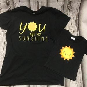 May include: Two black t-shirts with a yellow sun graphic. The larger shirt says "You are my sunshine".