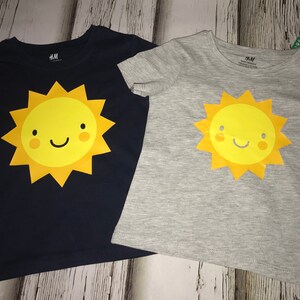May include: Two short-sleeved t-shirts featuring a smiling sun graphic. One shirt is navy blue and the other is light grey. Both shirts have the text "H&M" on the neckline.