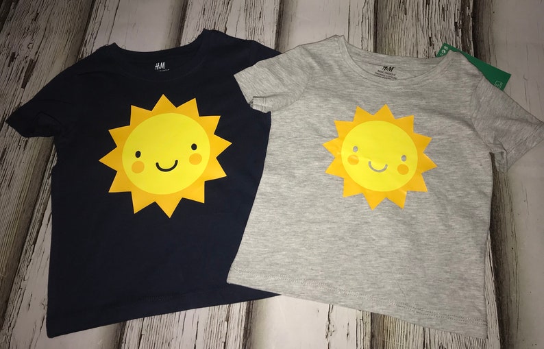 May include: Two short-sleeved t-shirts featuring a smiling sun graphic. One shirt is navy blue and the other is light grey. Both shirts have the text "H&M" on the neckline.