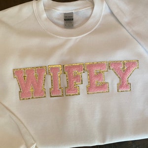 May include: White sweatshirt with the word "WIFEY" in pink and gold letters. The letters have a fuzzy texture and a gold outline.