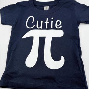 May include: A navy blue short-sleeved t-shirt with the text "Cutie Pi" in white. The Pi symbol is large and stylized.