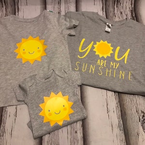 May include: Three grey t-shirts with a yellow sun graphic. The largest shirt says "You are my sunshine" with a yellow sun graphic.