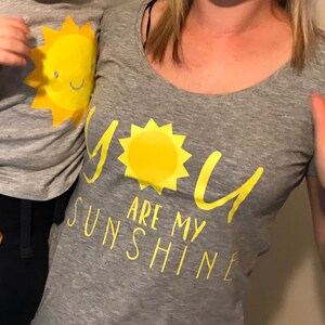 May include: A grey t-shirt with the text "You are my sunshine" in yellow. A yellow sun graphic is above the text. A child is wearing a grey t-shirt with a yellow sun graphic.