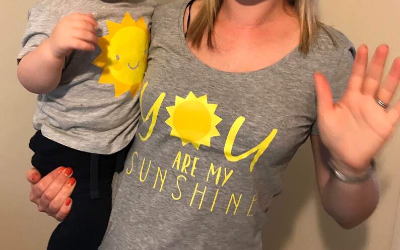 May include: A grey t-shirt with the text "You are my sunshine" in yellow. A yellow sun graphic is above the text. A child is wearing a grey t-shirt with a yellow sun graphic.