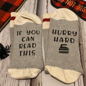 Puede incluir: Un par de calcetines grises con puños blancos y rayas rojas. Un calcetín dice "If you can read this" y el otro dice "Hurry hard" con un gráfico de piedra de curling.
