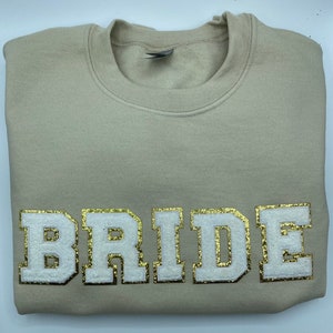 May include: A beige crewneck sweatshirt featuring the word BRIDE in large, block-style letters across the chest. The letters are white with a textured, glittery gold outline, creating a bold contrast against the solid light-coloured fabric.