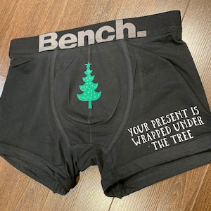 Puede incluir: Bóxer negro con el logo Bench gris en la cintura. Un gráfico de árbol de Navidad verde con puntos rojos está impreso en la parte delantera. El texto "YOUR PRESENT IS WRAPPED UNDER THE TREE" está impreso en el lado derecho.