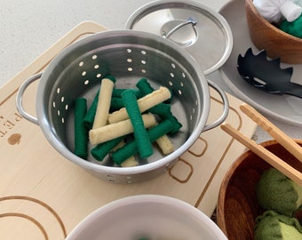 May include: A set of toy kitchen utensils and food, including a stainless steel pot with a strainer, a wooden cutting board, a wooden spoon, a pair of wooden tongs, a wooden bowl with green felt balls, and a white bowl with green and yellow felt balls.