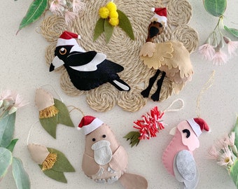 May include: A collection of felt bird ornaments, including a magpie, emu, and pink cockatoo, each wearing a Santa hat. Additional ornaments include leaf and flower designs. The ornaments are arranged on a woven placemat with eucalyptus sprigs.