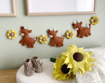 May include: A garland with brown felt highland cow figures and yellow felt flowers. The garland is hanging on a light green wall. There are two felt sunflowers on a white surface.