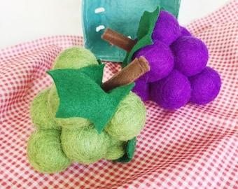 May include: Two bunches of felt grapes, one green and one purple, with felt stems and leaves. The grapes are sitting on a red and white checkered tablecloth.