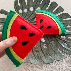May include: Two felt watermelon slices with red centers, black seeds, and green and yellow rinds. The slices are placed on a decorative leaf. The watermelon slices are handmade.