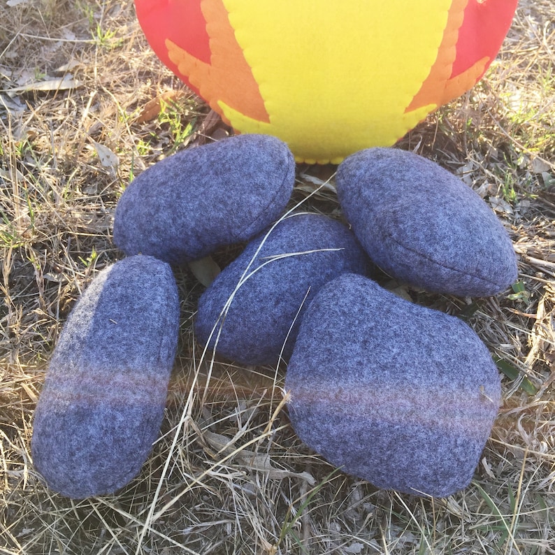 May include: Five gray felt rocks, resembling campfire stones, are arranged on a bed of dry grass.