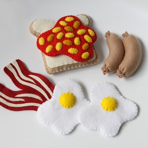 May include: Felt food set featuring a slice of toast with red jelly and yellow sprinkles, two fried eggs with yellow yolks, two brown sausages, and three strips of bacon.