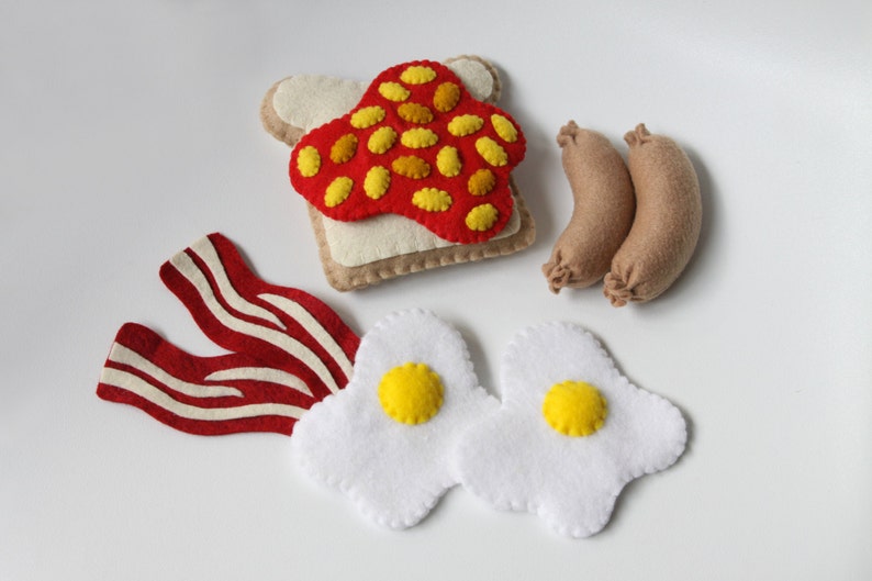 May include: Felt food set featuring a slice of toast with red jelly and yellow sprinkles, two fried eggs with yellow yolks, two brown sausages, and three strips of bacon.