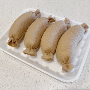 May include: Four brown felt sausages in a white styrofoam tray. The sausages are arranged in a row, with the ends of each sausage facing the viewer.