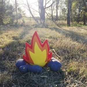 May include: A felt campfire with orange and yellow flames, surrounded by gray rocks. The campfire is set against a backdrop of a grassy field and trees.