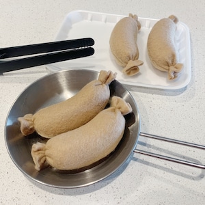 May include: Three brown felt sausages in a silver frying pan with a black handle. Two more brown felt sausages are on a white tray.