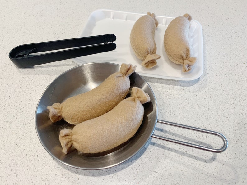 May include: Three brown felt sausages in a silver frying pan with a black handle. Two more brown felt sausages are on a white tray.
