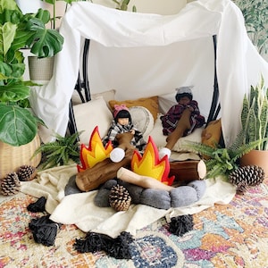 May include: Two dolls are sitting in a teepee made of white fabric. The dolls are wearing clothes and have long hair. There are two felt fire pit decorations in front of the dolls. The fire pit decorations are red, orange, and yellow.