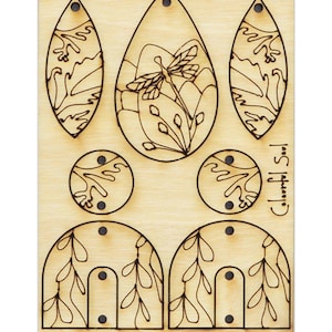 Könnte beinhalten: Eine Holzplatte mit sechs ausgeschnittenen Formen zum Herstellen von Ohrringen. Die Formen umfassen eine Träne mit einem Libellenmuster, ein Blatt und einen Halbkreis mit einem Blattdesign. Das Holz ist hellbraun und die Designs sind schwarz.