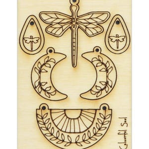 Könnte beinhalten: Holzplatte mit fünf lasergeschnittenen Designs für Ohrringe. Die Designs umfassen eine Libelle, zwei Halbmonde mit Blättern und zwei Tropfenformen mit Libellen. Die Platte ist mit "Colorful Soul" beschriftet.
