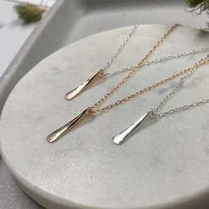 May include: Three delicate necklaces with simple, hammered pendant bars in silver, gold, and rose gold. The necklaces are displayed on a white marble surface.