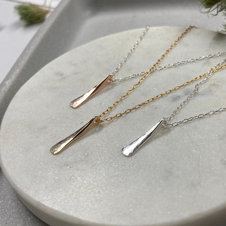 May include: Three delicate necklaces with simple, hammered pendant bars in silver, gold, and rose gold. The necklaces are displayed on a white marble surface.