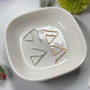 May include: Four triangle-shaped earring hooks in a white ceramic dish. Two hooks are silver and two are gold.