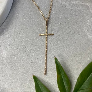 Pu&ograve; includere: Una collana con un ciondolo a forma di croce martellata in oro, su una catena delicata. La croce &egrave; lunga e sottile, con una superficie testurizzata.