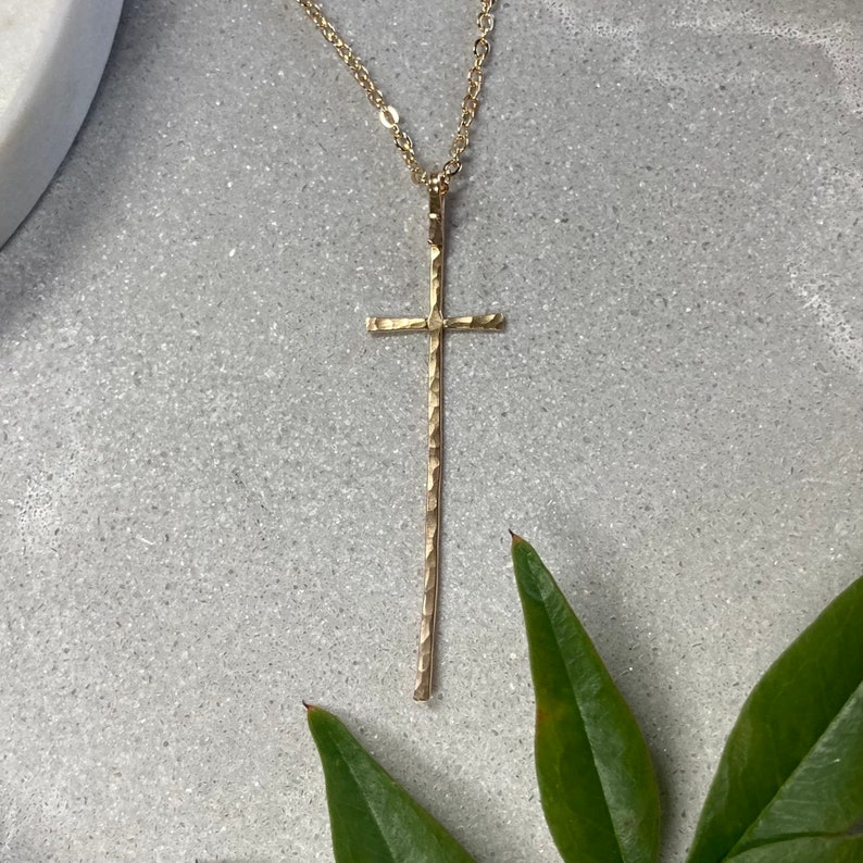 Pu&ograve; includere: Una collana con un ciondolo a forma di croce martellata in oro, su una catena delicata. La croce &egrave; lunga e sottile, con una superficie testurizzata.