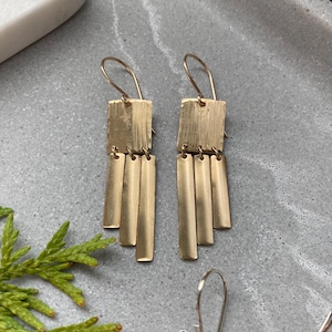 May include: Gold dangle earrings with a square top and four rectangular charms hanging below. The earrings have a brushed finish.