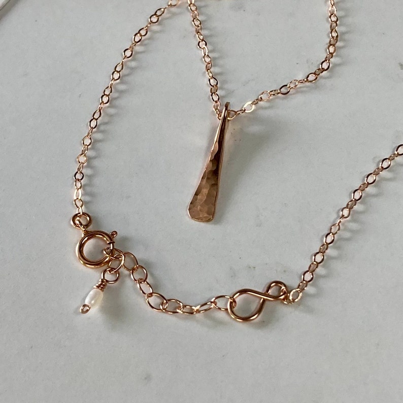 May include: A delicate rose gold necklace with a hammered pendant and a small pearl accent on the clasp.