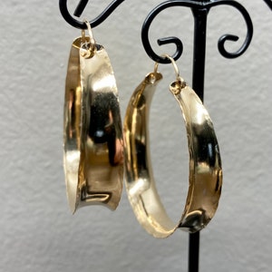 May include: A pair of large, gold-toned hoop earrings with a hammered finish. The earrings are slightly curved and have a wide, flat surface.