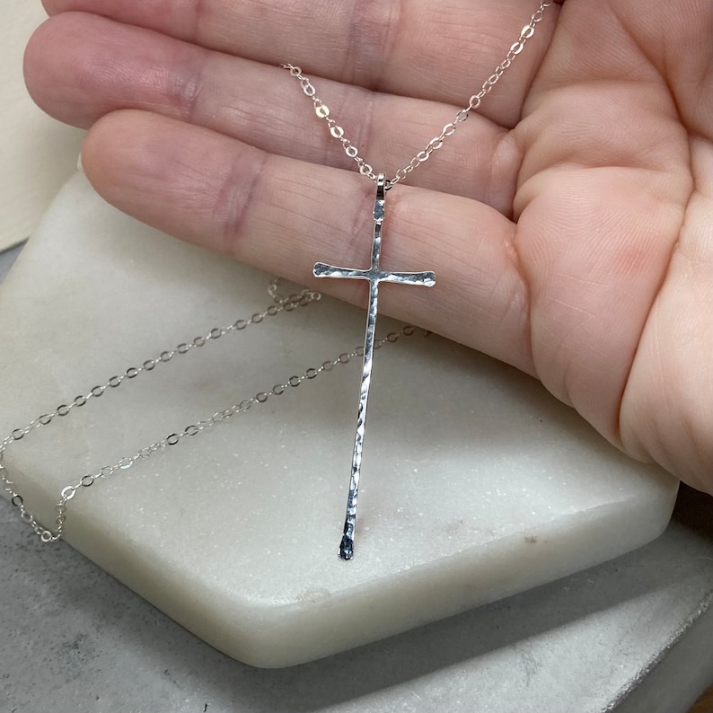 Pu&ograve; includere: Una collana d'argento con un ciondolo a forma di croce martellata. La catena &egrave; delicata e la croce &egrave; lunga e sottile.