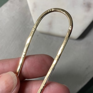May include: A gold-colored hairpin with a U-shaped design. The hairpin has a hammered texture and decorative lines near the top and bottom. The hairpin is held in a person's hand.