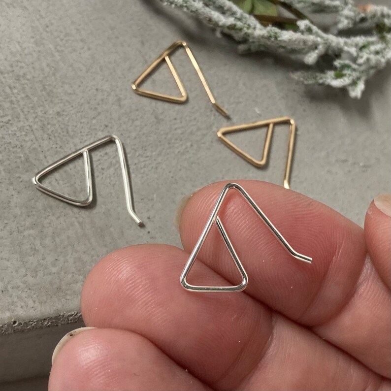 May include: A set of three triangle shaped earrings, two silver and one gold. The earrings are simple and minimalist in design.