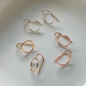 May include: A collection of six minimalist earrings in gold, silver, and rose gold tones. Each earring features a simple, open circle design with a curved post. The earrings are displayed on a white surface.