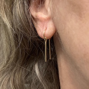 May include: Gold-colored, minimalist earrings. Each earring features a small, curved hook that attaches to the ear, with two vertical, rectangular bars hanging below. The earrings are simple and elegant, suitable for everyday wear.