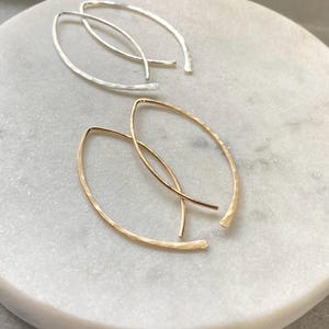 May include: Two pairs of minimalist earrings. One pair is silver, the other is gold. Both pairs have a hammered texture and a curved, elongated shape. The earrings are displayed on a white and gray marble surface.