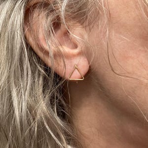 May include: A close-up of a gold-colored triangle stud earring. The earring is a simple geometric design, a hollow triangle shape. The earring is worn on a person's ear, with the focus on the earring's design and color.